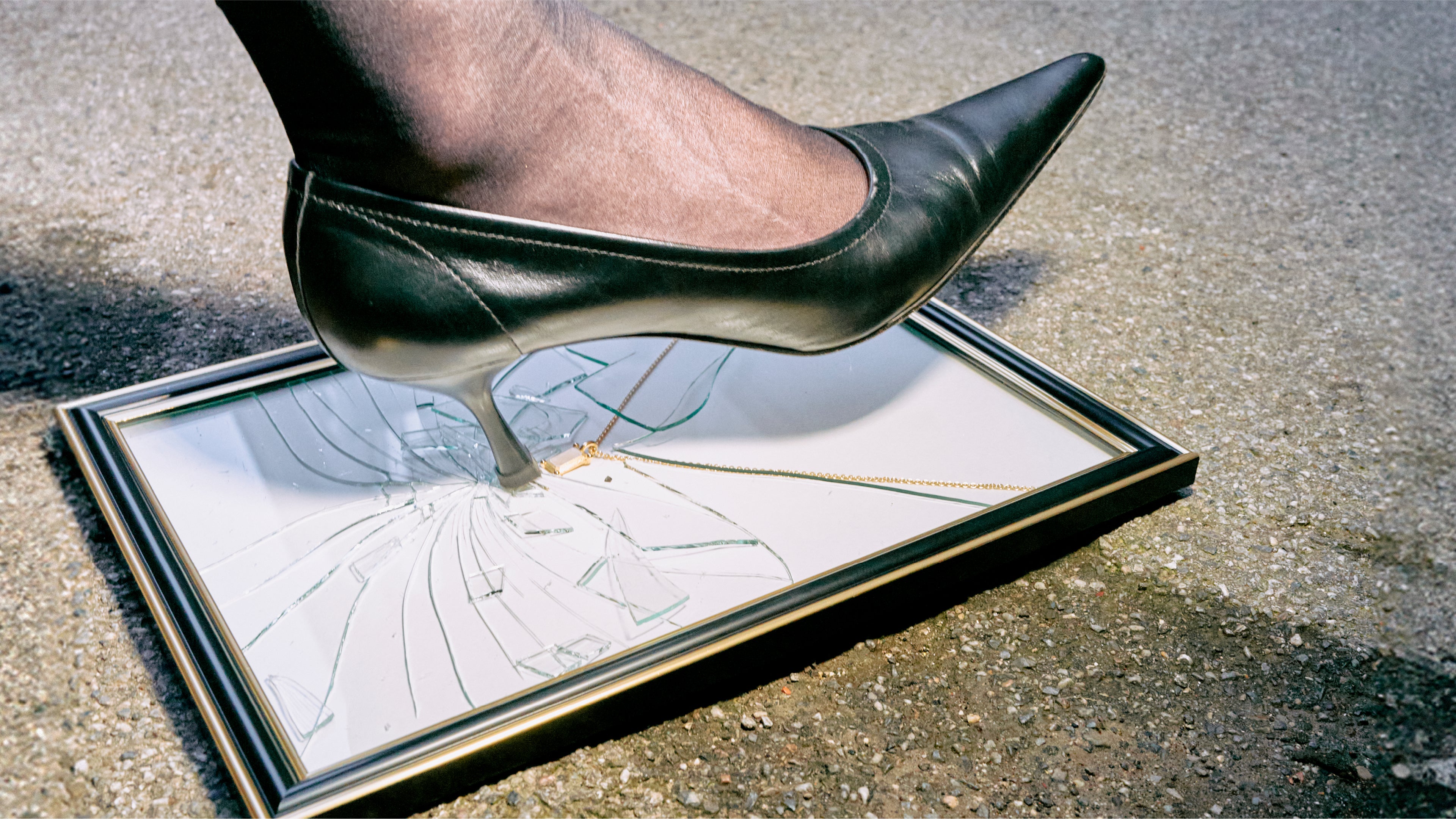 A picture frame lies on the floor containing an emerald charm on an oval chain. A foot wearing a stiletto heel steps on the glass of the frame, causing it to shatter.